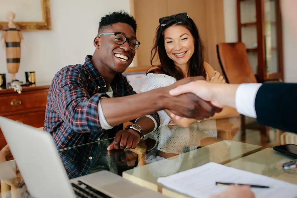 Homebuyers shaking hands with a loan officer across a desk with paperwork and a laptop