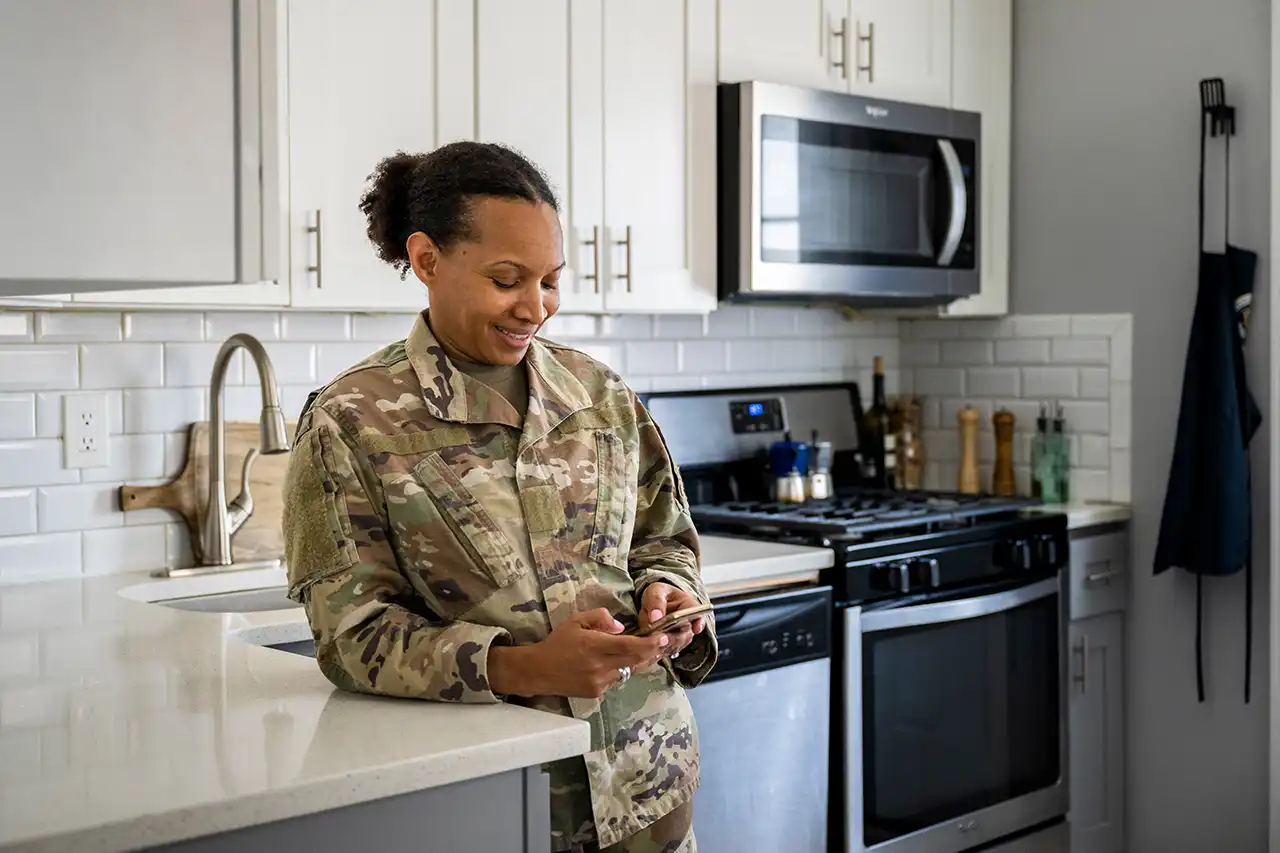 Service member in uniform reviewing information on a phone in a kitchen, representing early VA loan planning steps